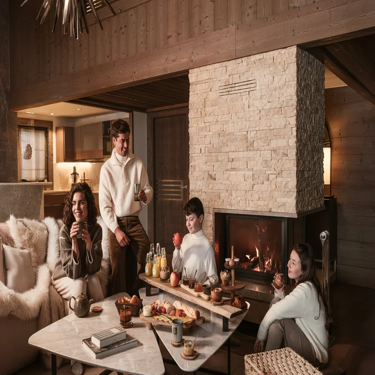 Guests gather in a serene lounge with a stone fireplace, sharing pastries, fruit, and beverages at a central table in an inviting setting.