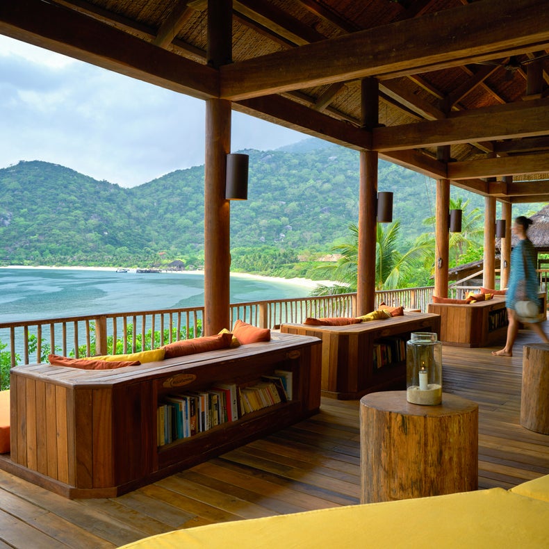 Salon en bois doté de sièges rembourrés et de bibliothèques, avec vue sur la montagne, la plage et l'océan à travers de larges ouvertures sous un ciel nuageux.