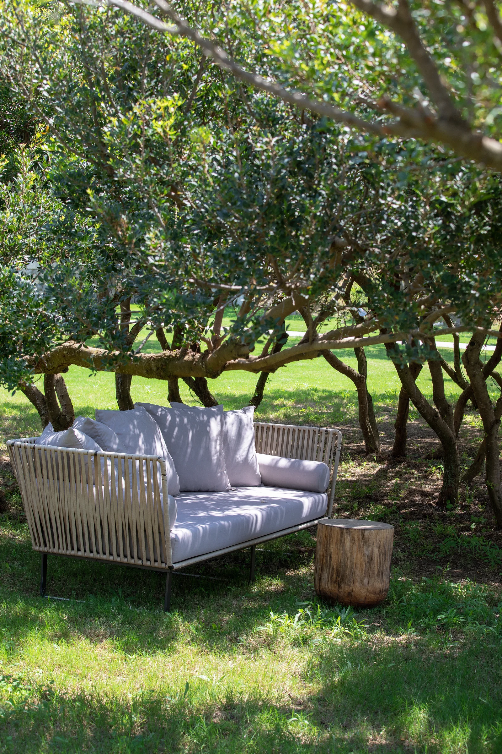 A woven outdoor sofa placed under old olive trees on cut grass, a log side table beside it and low branches shading the seat.