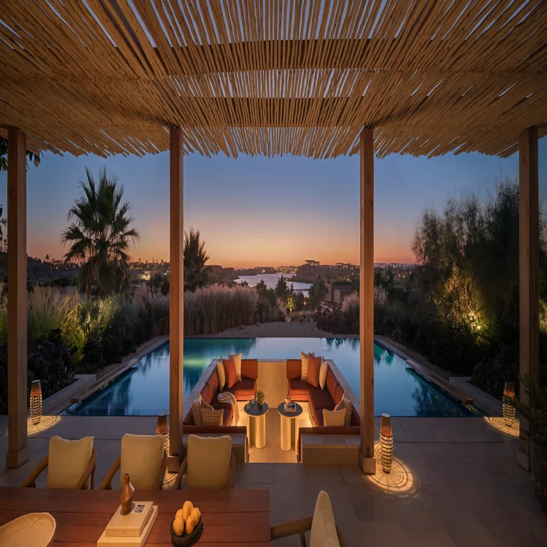 A sunken seating area by a pool features views of the sunset, distant water, and landscape, surrounded by a wooden pergola and abundant greenery.