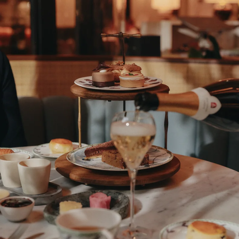 Sparkling wine is poured into a glass next to trays of pastries, cakes, and scones on a marble table prepared for afternoon tea in a softly lit café.