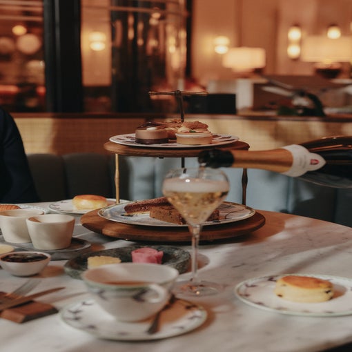 Sparkling wine is poured into a glass next to trays of pastries, cakes, and scones on a marble table prepared for afternoon tea in a softly lit café.