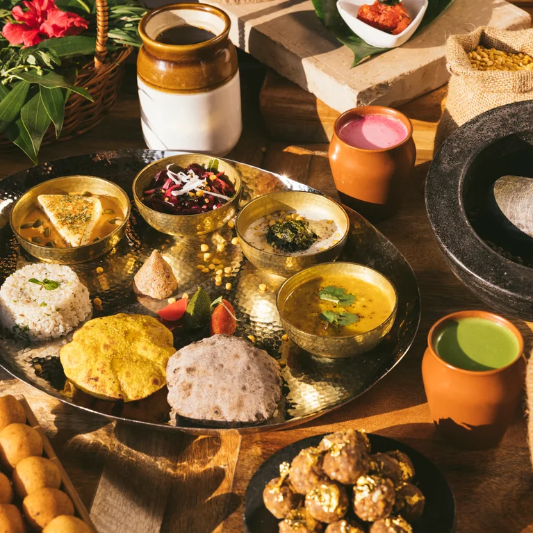 各种印度咖喱、米饭、面包、甜点、饮料和香料摆放在木桌上,阳光照射下,新鲜的食材显得格外耀眼。.