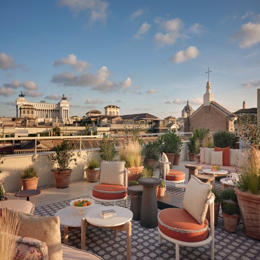 Outdoor area featuring round tables and cushioned chairs in orange and white, surrounded by potted greenery with city views beneath a partly cloudy sky.