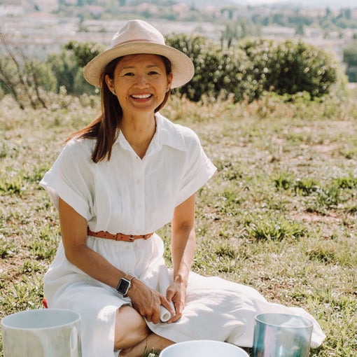 A person outdoors sits on grass with crystal singing bowls arranged before them, surrounding landscape visible in the background.