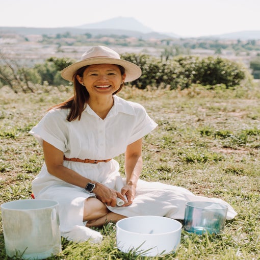 A person outdoors sits on grass with crystal singing bowls arranged before them, surrounding landscape visible in the background.