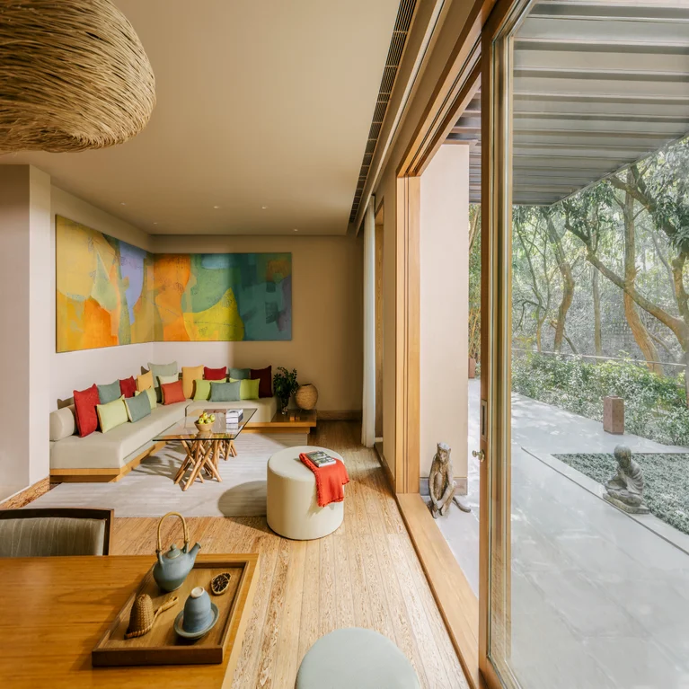 Living space with expansive windows, light wood floors, abstract art, beige sectional sofa with vibrant pillows, overlooking a tranquil green garden.