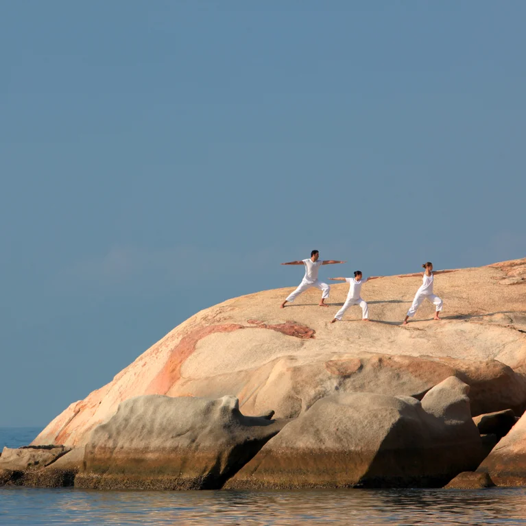 يقوم الضيوف الذين يرتدون ملابس بيضاء بممارسة اليوغا على الصخور الساحلية الملساء بجانب البحر، حيث تغمرهم أشعة الشمس الطبيعية والسماء الصافية في الأعلى.
