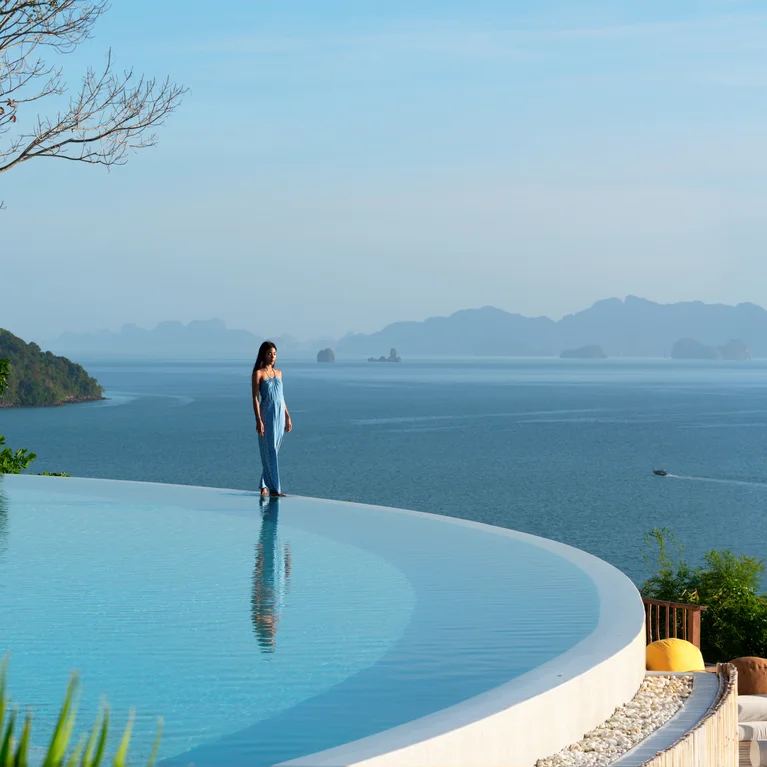 一位客人在无边泳池边驻足,泳池面朝宁静的大海,背景是晴空下清晰可见的岛屿和青山。.