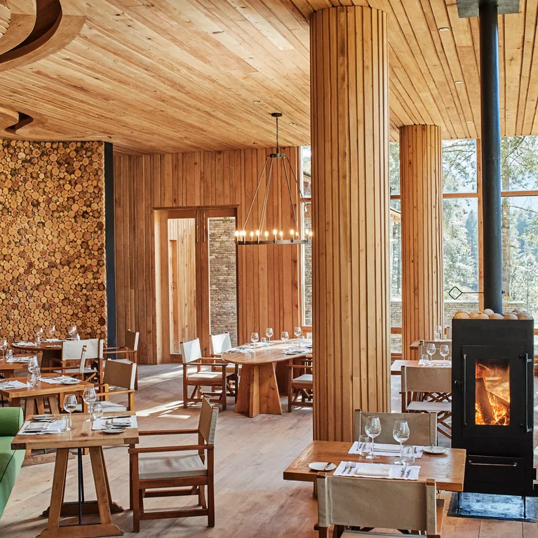 餐厅的墙壁和天花板采用木质镶板,细节处采用天然木料,配有绿色沙发、套桌、大窗户和燃木火炉。.