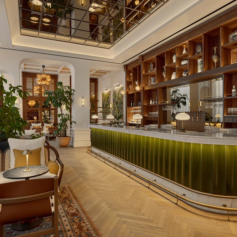 Bar with a green counter, natural wood shelving holding bottles, comfortable chairs, lush indoor plants, and inviting soft lighting designed for relaxation and sustainability.