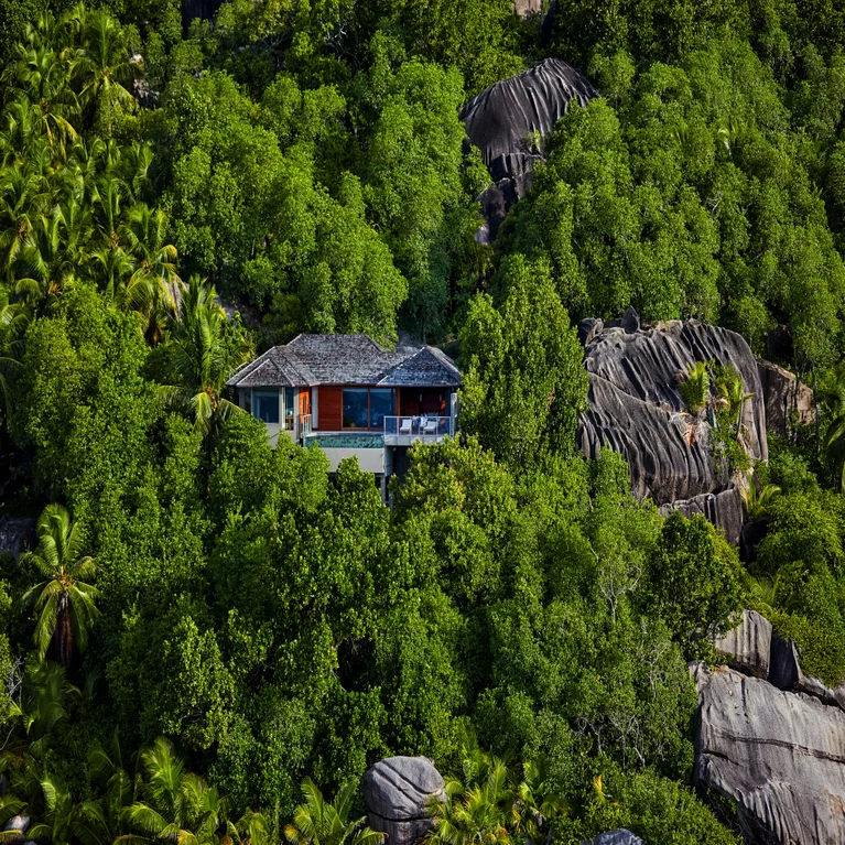 A wooden-roofed residence surrounded by green trees and granite rocks rests on a hillside, thoughtfully integrated into its natural environment.
