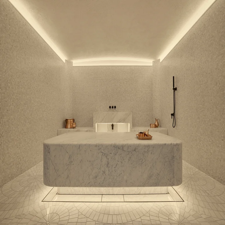 Hammam room with a central marble slab, gentle lighting, mosaic tile walls and floor, copper bowls for rituals, and an accessible shower fixture on the right.
