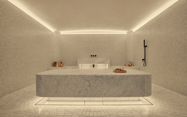 Hammam room with a central marble slab, gentle lighting, mosaic tile walls and floor, copper bowls for rituals, and an accessible shower fixture on the right.