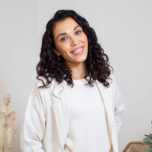 Person in a light blazer and white shirt stands indoors by greenery and sculptural decor, set against a soft-toned backdrop. The scene highlights natural elements.