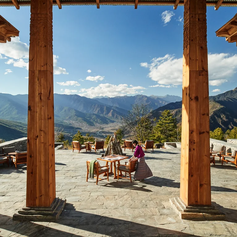 Terrace with wooden pillars, mountain views and natural light; individual in traditional clothes stands near a table, under a bright sky.