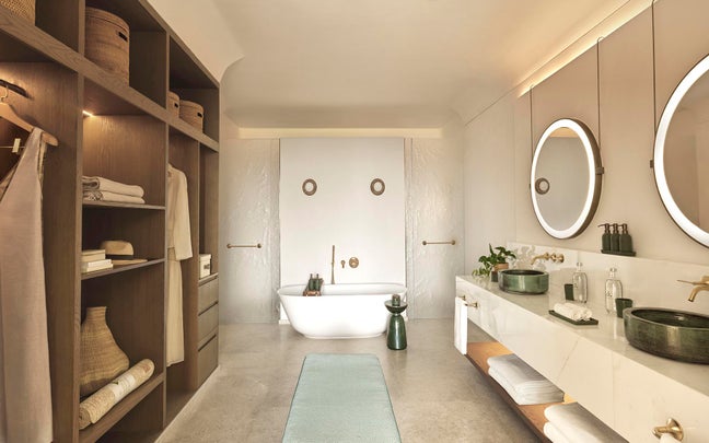 Bathroom featuring a freestanding tub, double sinks, round illuminated mirrors, open shelves, and neutral hues accented by green elements.