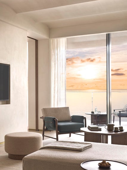 A serene living area with a comfortable lounge chair and table faces calm sea views and an orange sunset through expansive glass windows.