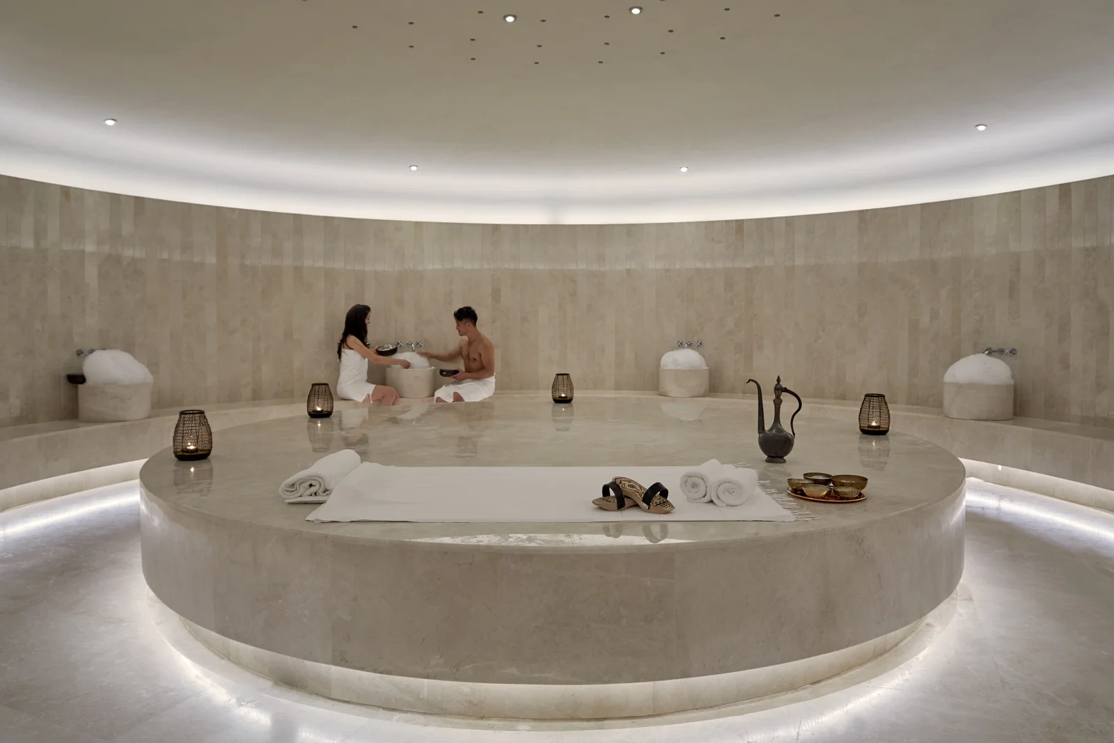 The hammam, a circular marble slab lit from beneath in a domed stone room, with two guests seated at the far side and brass vessels around the rim.