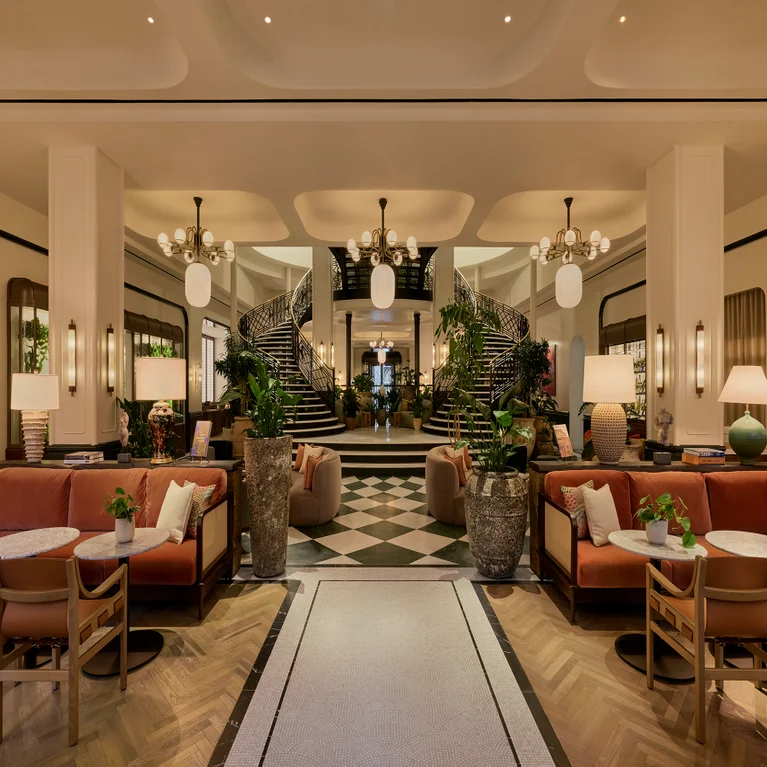Lobby with natural wood and black-and-white flooring, eco-friendly seating, abundant greenery, and a wide double staircase in softly lit surroundings.
