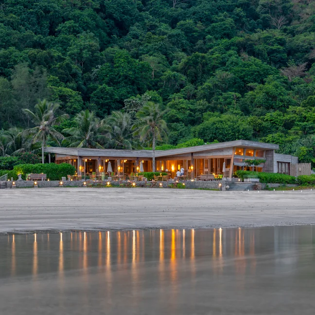 黄昏时分,一幢拥有宽大窗户的海滨别墅在葱郁树木的映衬下,发出柔和的灯光,在潮湿的沙滩上映出别墅的身影。.