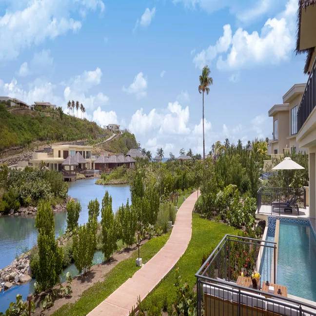 Pathway leads alongside a river bordered by greenery, with contemporary villas and a pool visible, set beneath a blue sky with gentle clouds.