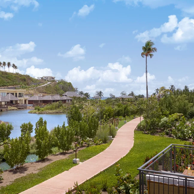 Pathway leads alongside a river bordered by greenery, with contemporary villas and a pool visible, set beneath a blue sky with gentle clouds.