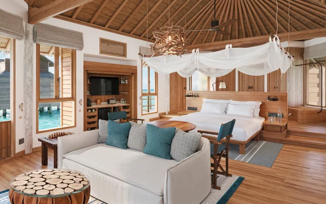 A bedroom in a villa with a bed, seating area, and open wooden doors looking out toward the ocean.
