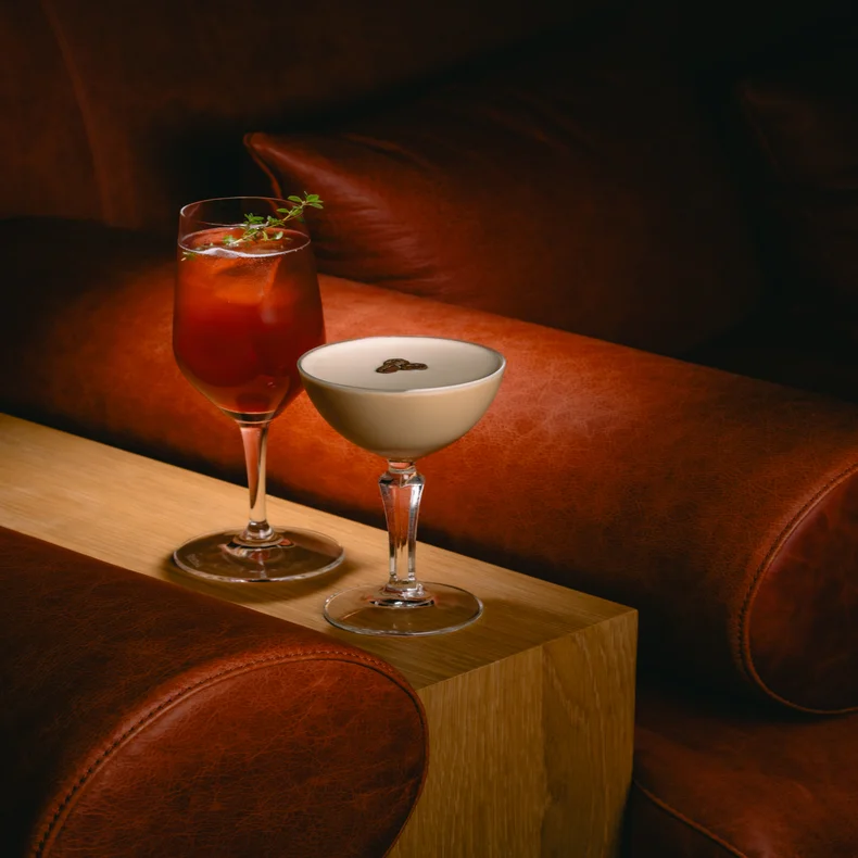 コーヒー豆をトッピングしたクリーミーなコーヒードリンクの横に、氷とガーニッシュを添えた赤いカクテルが、茶色の革張りの席の前に置かれた木の上に置かれている。.