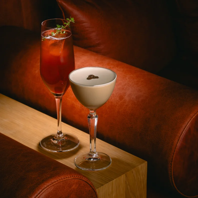 コーヒー豆をトッピングしたクリーミーなコーヒードリンクの横に、氷とガーニッシュを添えた赤いカクテルが、茶色の革張りの席の前に置かれた木の上に置かれている。.