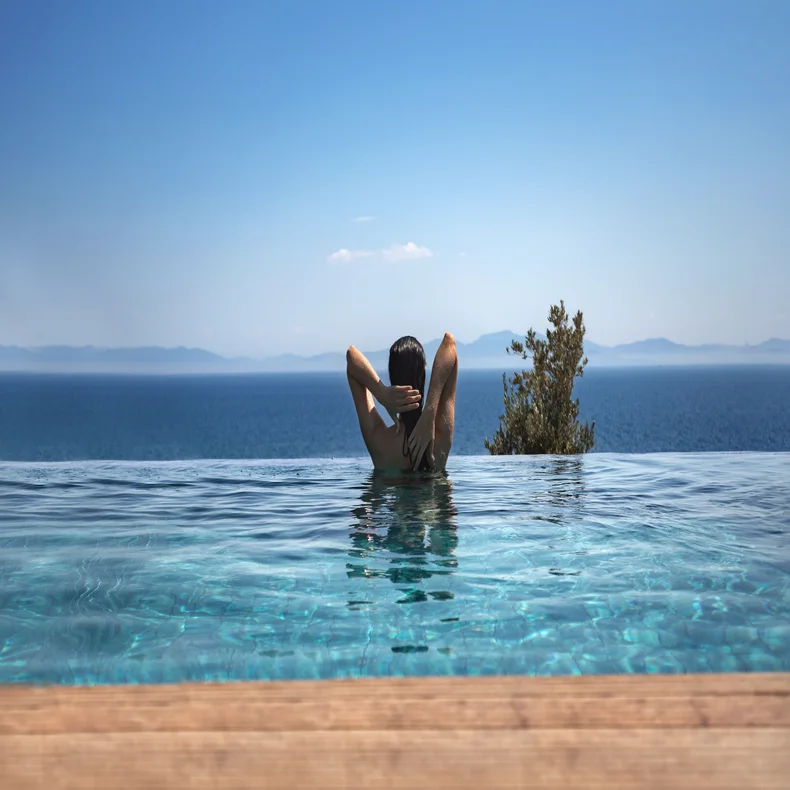 Guest relaxes in an infinity pool overlooking a tranquil blue sea, distant mountains and open sky, hands gently placed behind their head.