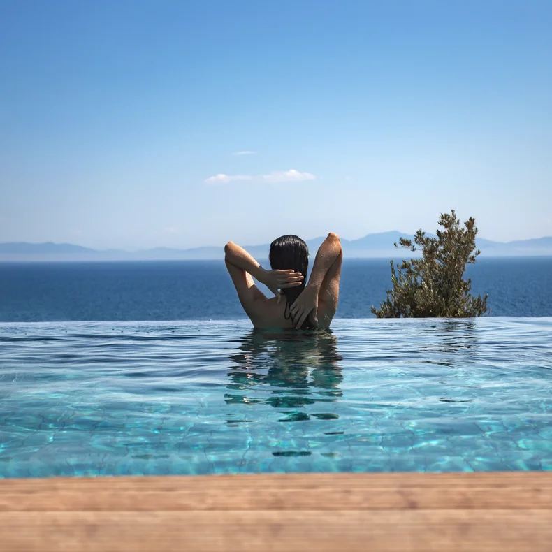 Guest relaxes in an infinity pool overlooking a tranquil blue sea, distant mountains and open sky, hands gently placed behind their head.