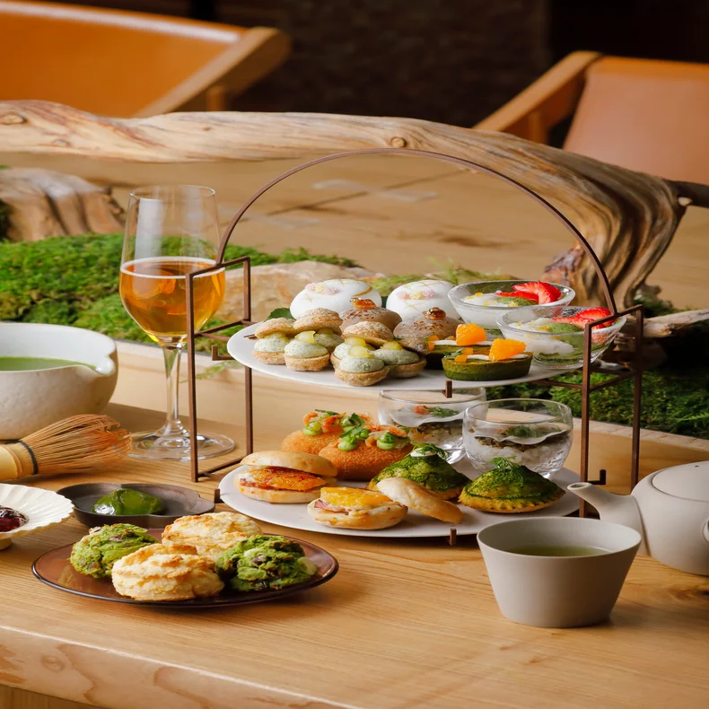 Delicately arranged pastries and savory selections, served with tea, sparkling beverage and scones on a wooden table beside fresh greenery.
