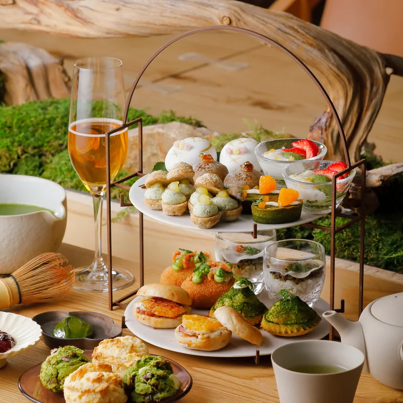 繊細にアレンジされたペストリーとセイボリーセレクションを、紅茶、スパークリングドリンク、スコーンとともに、新緑に囲まれた木製のテーブルで。.