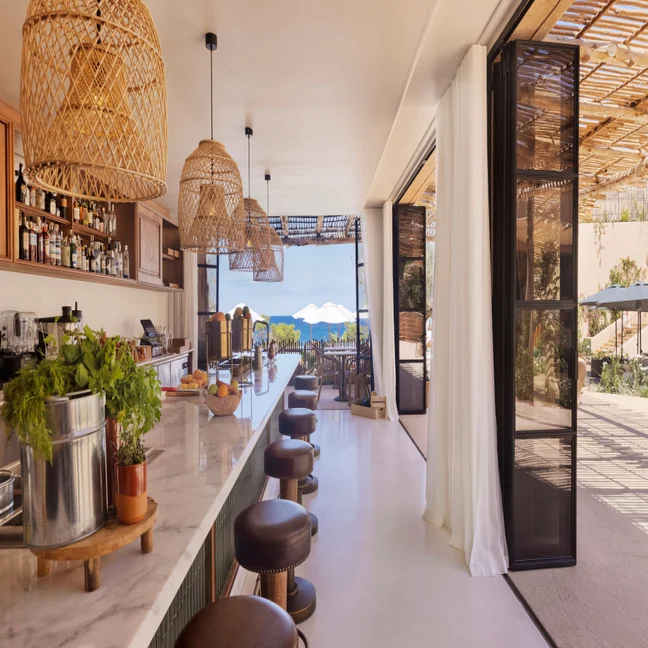 Bar featuring a marble counter and wicker pendant lights, with open doors to a patio shaded by umbrellas and views of the ocean in the distance.