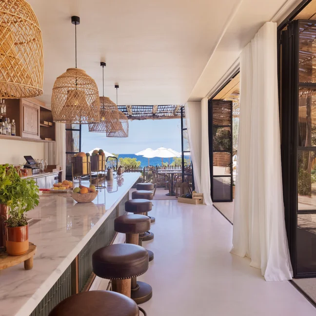 Bar featuring a marble counter and wicker pendant lights, with open doors to a patio shaded by umbrellas and views of the ocean in the distance.