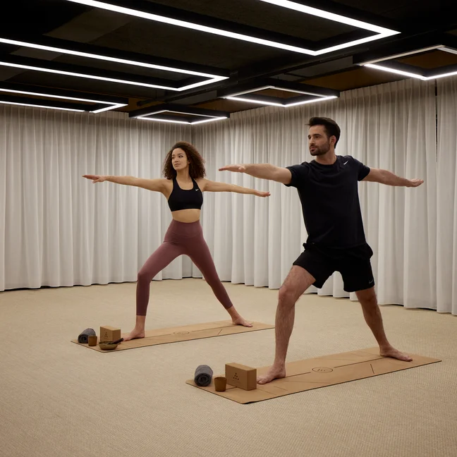 Guests hold a wide-legged yoga pose with extended arms on mats in a bright studio. Accessible props, including blocks and towels, are placed within easy reach.