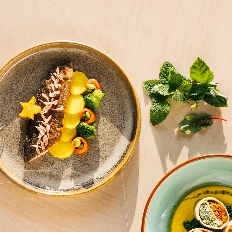 烤鱼配黄色蔬菜、西兰花和胡萝卜片放在叉子旁边;新鲜香草和另一种蔬菜放在浅色桌面上。.