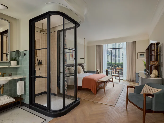 Thoughtfully designed room featuring a glass shower, bed with orange accents, sitting area, expansive windows, and natural-toned decor.