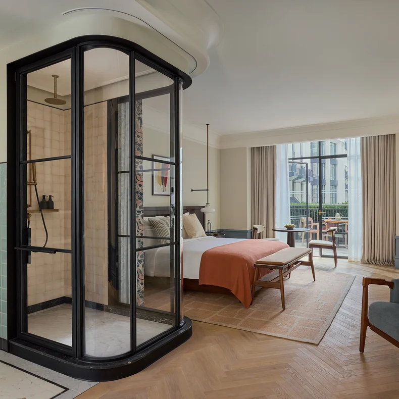 Thoughtfully designed room featuring a glass shower, bed with orange accents, sitting area, expansive windows, and natural-toned decor.