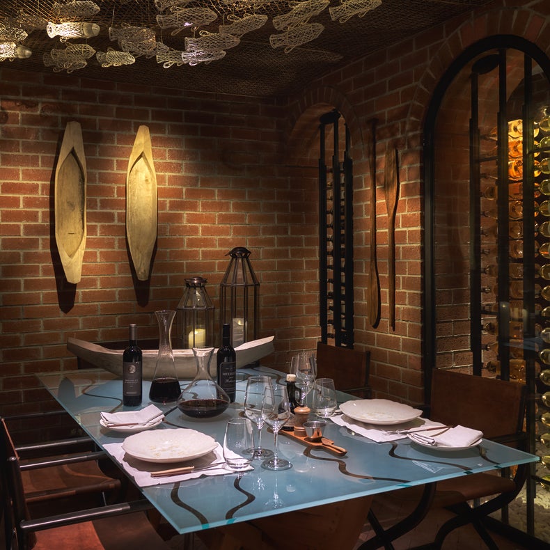 Zona de comedor con paredes de ladrillo visto, una mesa de cristal para seis personas, remos decorativos en la pared, botellas de vino y una suave iluminación ambiental.