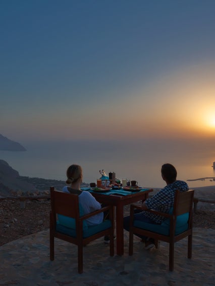 Los huéspedes comparten una comida en una mesa al aire libre en una terraza de montaña, con vistas al mar y a la costa mientras la cálida luz natural llena el cielo del atardecer.
