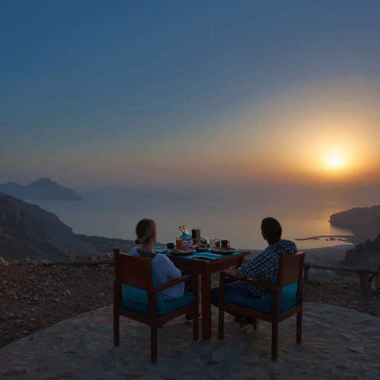 Los huéspedes comparten una comida en una mesa al aire libre en una terraza de montaña, con vistas al mar y a la costa mientras la cálida luz natural llena el cielo del atardecer.