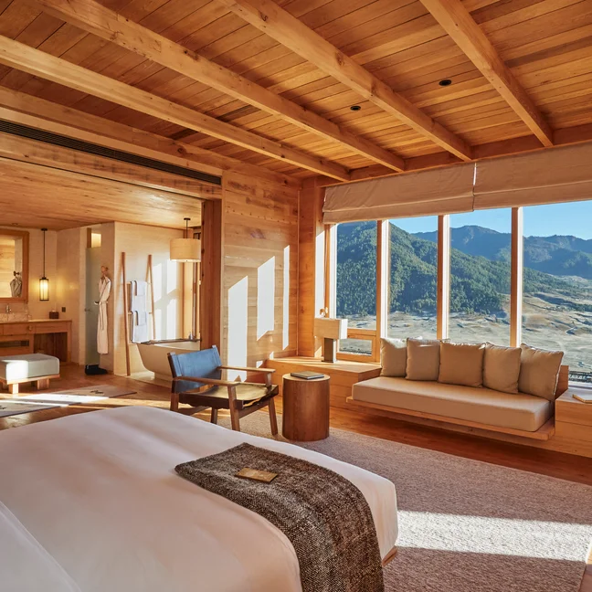 木质装饰的客房拥有宽大的窗户、床、沙发和山景。在明亮的自然光线下,一位身着白色睡袍的客人正在浴室附近。.