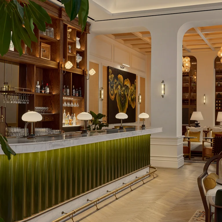 Inviting bar area featuring a green and white counter, refined lighting, wooden shelves holding bottles, comfortable seating, and abundant indoor greenery.