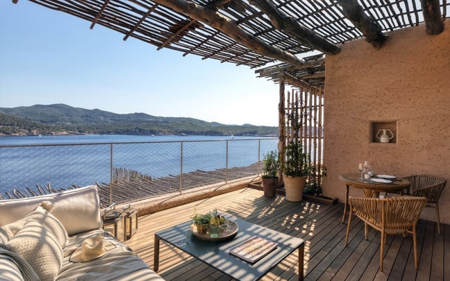 A suite terrace under a reed-slatted pergola, a cushioned daybed and low table facing a rope balustrade with open sea beyond, and a small table set for two against the rendered wall.