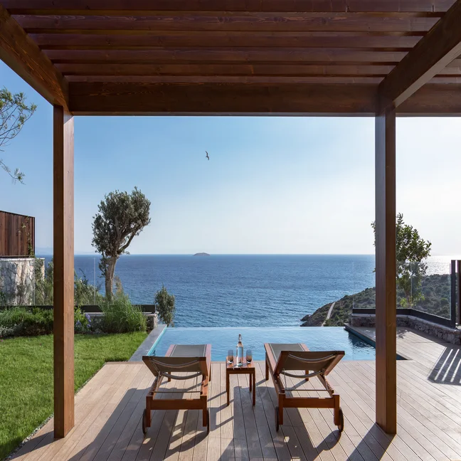 A peaceful outdoor setting features an infinity pool, ocean view, shaded seating under a wooden pergola, and clear sky extending to the horizon.