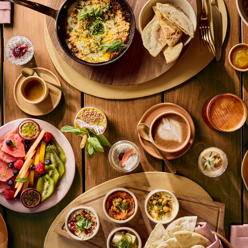 Breakfast selection featuring pastries, fruits, eggs, dips, coffee and juice arranged on wooden and pink plates, viewed from above.