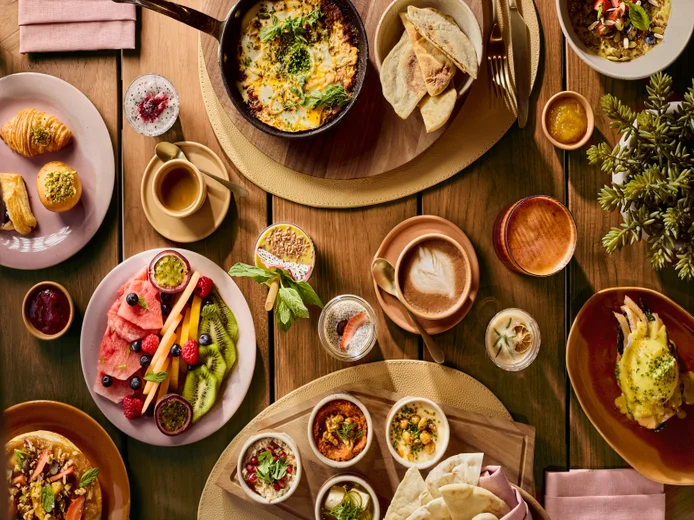 Breakfast selection featuring pastries, fruits, eggs, dips, coffee and juice arranged on wooden and pink plates, viewed from above.
