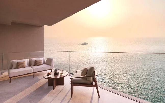 Balcony seating with a sofa, chair and table faces the sea at sunset; soft sunlight fills the space and a distant boat crosses calm water.
